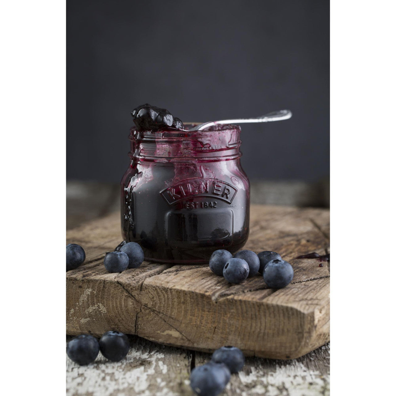 Jar of blueberry jam with a spoon on a wooden board, surrounded by blueberries.