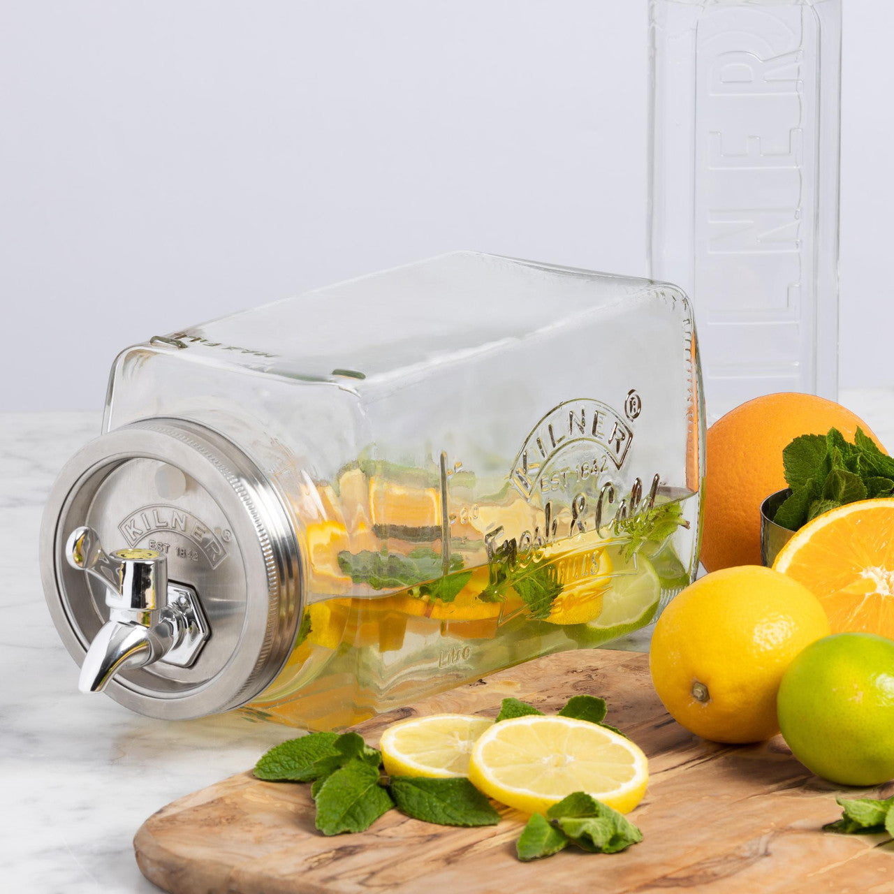 Clear glass carafe with metal spigot on a wooden cutting board with lemons and oranges.