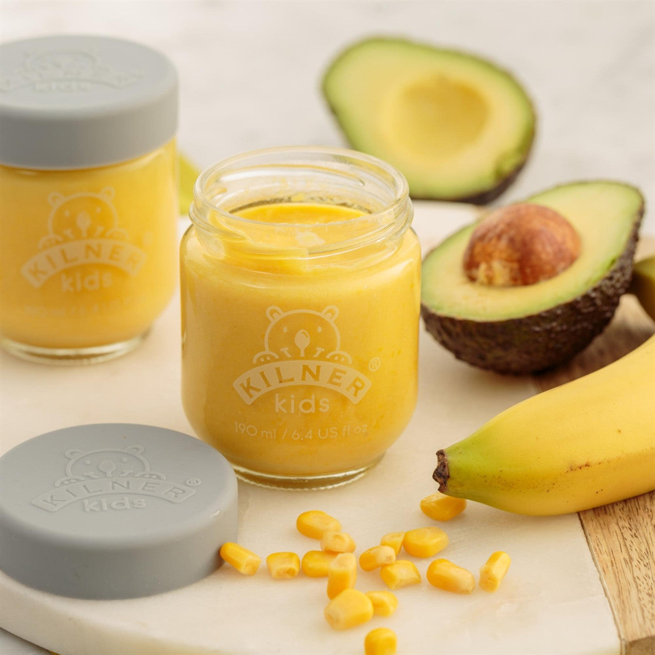 Two jars of yellow baby food with a halved avocado and banana on a light surface.