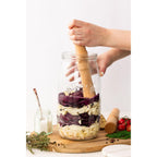Glass jar with layered ingredients on a wooden board, being pressed by a wooden tool.