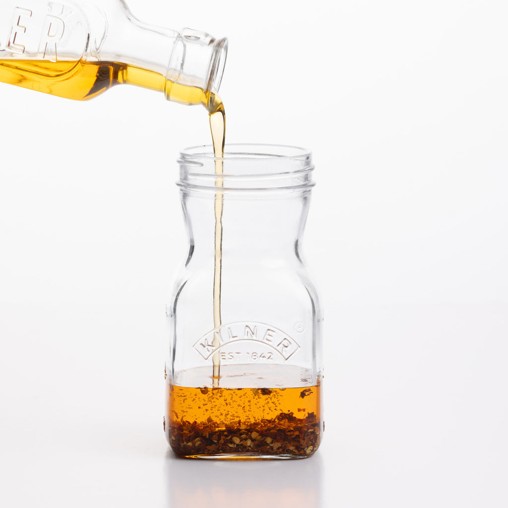Oil being poured from a glass bottle into a glass jar with a brand logo on a white background