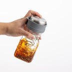 Hand holding a glass jar with a metal lid filled with a mixture of ingredients on a white background