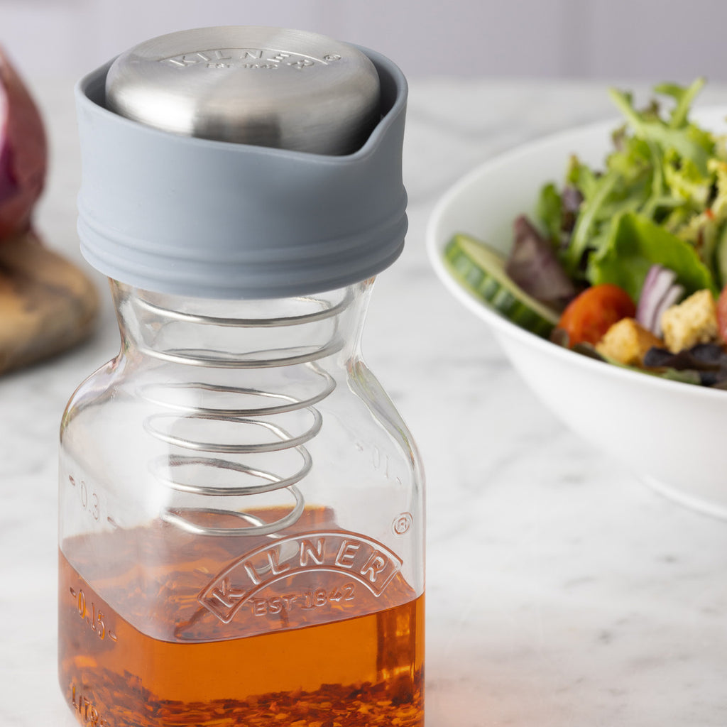 Glass bottle with a gray cap containing a liquid, placed on a surface with a blurred salad in the background.