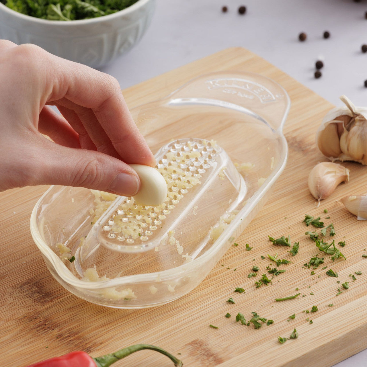 Hand using a garlic press on a wooden cutting board with chopped garlic and herbs.