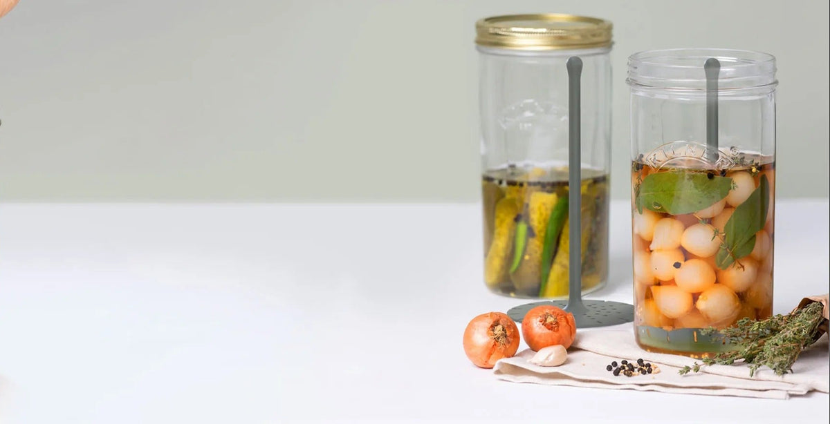 A hand is shown adding ingredients into a clear glass jar with amber liquid, next to two finished pickled or preserved jars on a white surface against a light gray background.